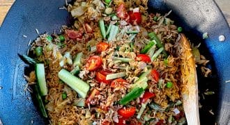 A wok filled with fried rice mixed with peas, tomatoes, cucumber slices, greens, and minced meat, with a wooden spoon resting on the side. The dish sits on a wooden table.