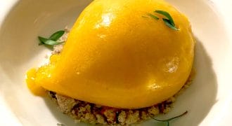 A bright yellow scoop of mango sorbet sits on a bed of crumbled biscuit, garnished with a few small green herb leaves, served in a white bowl.