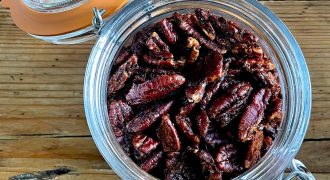 A glass jar filled with roasted pecan halves sits open on a rustic wooden surface, with the orange-rimmed lid resting to the side and attached by a metal clasp.
