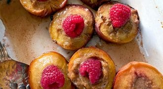 Halved baked peaches topped with a dollop of brown filling and fresh raspberries, arranged in a white baking dish with a serving spoon at the side.