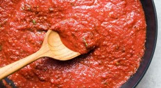 A wooden spoon stirs a thick, red tomato sauce with herbs in a black pan, viewed from above.