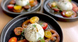 Five plates each feature a salad of sliced red and yellow heritage tomatoes, topped with a ball of burrata cheese, fresh herbs, black pepper, and a drizzle of olive oil, arranged on dark round plates.