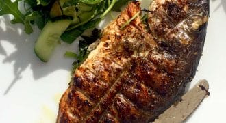Grilled whole fish served on a white plate with a side of fresh green salad, including rocket and cucumber, accompanied by a fork and a knife.