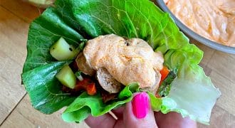 A hand with pink nail varnish holds a lettuce wrap filled with grilled chicken, diced vegetables, and topped with a dollop of orange creamy sauce. Bowls of salad and sauce are in the background on a wooden surface.