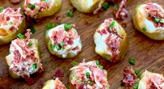 Bite-sized baked potato pieces topped with soured cream, crumbled bacon, and chopped chives are arranged on a wooden surface.