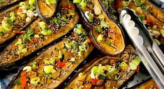 Roasted aubergine halves topped with chopped spring onions, red chilli, sesame seeds, and herbs are arranged on a tray lined with foil. Metal tongs are placed on the right side of the tray.