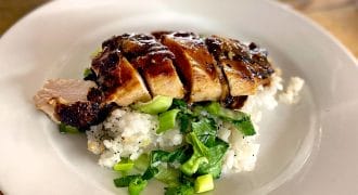 Sliced grilled chicken breast served on a bed of white rice and chopped green vegetables, arranged on a white plate on a wooden table. A blurred wine glass is visible in the background.