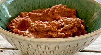 A decorative ceramic bowl filled with a chunky, reddish-brown dip sits on a wooden table, with a blurred outdoor scene in the background.