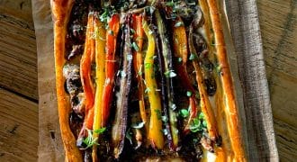 A rectangular vegetable tart with flaky golden pastry, topped with colourful roasted carrots, herbs, and a dark spread, sits on baking parchment on a wooden chopping board in sunlight.