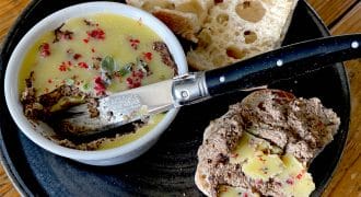 A black plate with a ramekin of pâté topped with herbs and spices, slices of rustic bread, and a knife spread with pâté and butter on a wooden table.