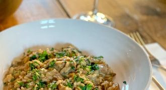 A white bowl of creamy mushroom risotto garnished with chopped parsley sits on a wooden table beside a glass of white wine, with a fork and serviette nearby.