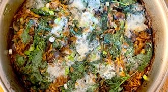 A pot of cooked orzo pasta mixed with spinach, mushrooms, spring onions, and melted cheese, sitting on a wooden table. The ingredients are combined and lightly browned on top.
