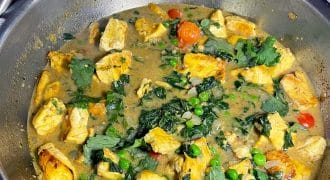 A stainless steel pan filled with a chicken curry dish, featuring chunks of chicken, peas, cherry tomatoes, fresh coriander, and a creamy sauce. A bowl of white rice is visible in the background.