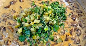 A creamy mushroom stew in a saucepan, topped with chopped gherkins and fresh parsley.