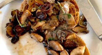 Two slices of toasted bread topped with sautéed mushrooms and herbs on a white plate, with a fork and knife placed beside the food.