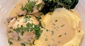 A plate with creamy mashed potatoes, chicken in herb sauce, and a serving of cooked dark leafy greens, all garnished with chopped fresh herbs.