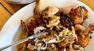 A platter of roasted, carved chicken with metal tongs, served on a wooden table next to a bowl of fresh green salad with broccoli and nuts.