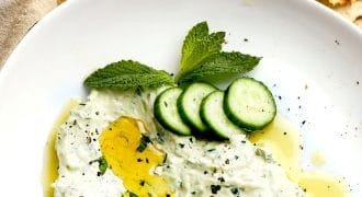 A white plate with creamy tzatziki dip, garnished with olive oil, mint leaves, and cucumber slices. Pieces of flatbread are in the background.