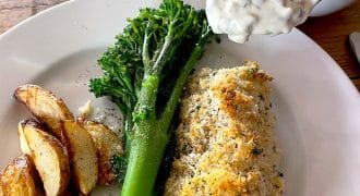 A plate with baked fish topped with breadcrumbs, roasted potato wedges, and steamed tenderstem broccoli. A spoonful of creamy tartare sauce is being served from a small bowl.