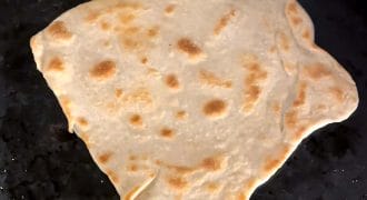 A piece of unevenly shaped flatbread with golden-brown spots cooks on a dark, oily griddle.