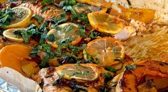 Baked salmon fillet topped with lemon slices and fresh herbs, surrounded by roasted orange and yellow vegetables on a foil-lined baking tray.