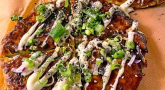 Two savoury Japanese pancakes (okonomiyaki) topped with mayonnaise, chopped spring onions, red onions, sesame seeds, and herbs, sitting on brown baking parchment.