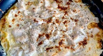 A black frying pan on a wooden table contains a cooked, golden-brown omelette topped with melted cheese and black pepper. The omelette appears fluffy with slightly crispy edges.