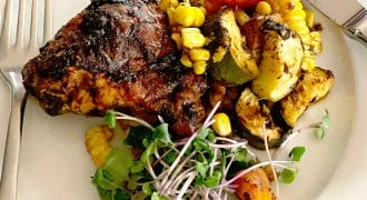 A plate with grilled chicken, roasted vegetables including courgette, tomato, and sweetcorn, garnished with fresh microgreens. A fork and knife are placed beside the food on the white plate.