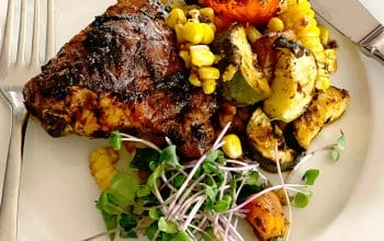 A plate with grilled chicken, roasted vegetables including courgette, tomato, and sweetcorn, garnished with fresh microgreens. A fork and knife are placed beside the food on the white plate.