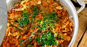 A large saucepan filled with a stew containing sausages, vegetables, orzo pasta, and chopped herbs on top. Flatbread and a glass are placed on the wooden table beside the saucepan.