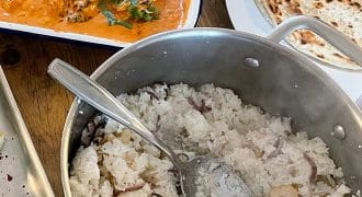A pot of cooked white rice with cashews and onions sits next to a dish of creamy orange curry garnished with coriander and a plate of round flatbread on a wooden table.