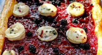 Rectangular puff pastry tart topped with a red jam-like layer, blackberries, and round slices of goats' cheese, garnished with herbs and pink peppercorns. The crust is golden and flaky.