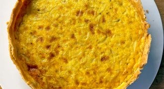 A golden-brown quiche with a flaky pastry sits on a white plate atop a wooden surface. The filling appears smooth and slightly browned on top.