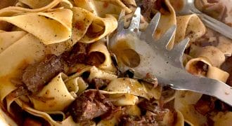 Wide ribbons of pasta tossed with chunks of braised beef in a rich, dark sauce, served in a white dish with a pasta server and metal spoon.