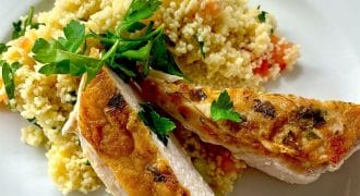 A white plate with grilled chicken breast slices garnished with parsley, served alongside a mound of couscous mixed with chopped vegetables.