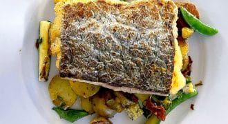 A fillet of fish with crispy skin served on sautéed vegetables, including courgette, potatoes, and green beans, on a white plate.