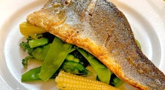 A cooked fish fillet with crispy skin served on a bed of baby corn, broccoli, and mangetout, presented on a white plate.