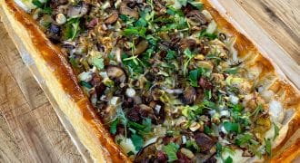 A rectangular puff pastry tart topped with sliced mushrooms, fresh herbs, and onions sits on baking parchment on a wooden surface. A knife is visible nearby.
