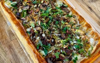 A rectangular puff pastry tart topped with sliced mushrooms, fresh herbs, and onions sits on baking parchment on a wooden surface. A knife is visible nearby.