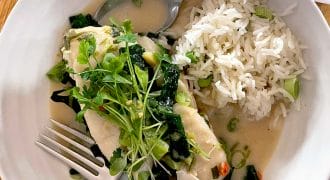 A white bowl with steamed white fish topped with fresh herbs and greens in a light sauce, served alongside a portion of white rice with spring onions. A fork and spoon rest on the edge of the bowl.