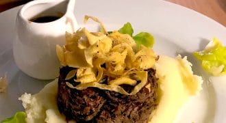 A plated dish featuring shredded beef topped with crispy fried onions, surrounded by creamy mashed potato and garnished with green leaves, with a small jug of brown sauce on the side.