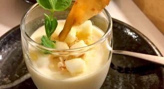 A glass dessert dish filled with creamy panna cotta, topped with diced fruit, a crunchy garnish, and a small green sprig, placed on a dark plate with a spoon beside it.