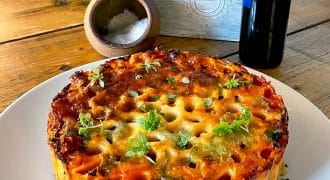A baked pasta pie with a golden, cheesy top and herbs is served on a white plate. There’s a wooden table, a salt pot, a bottle, and a planter with greens in the background.