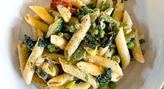 A white bowl filled with penne pasta mixed with green peas, leafy greens, and a creamy sauce, topped with cracked black pepper, sits on a wooden table.