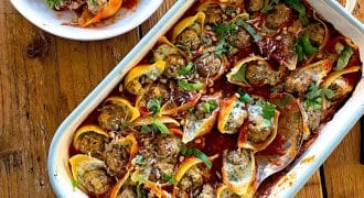 A baking dish filled with stuffed pasta shells topped with tomato sauce and herbs, with a serving spoon and a bowl of mixed green salad beside it on a wooden table.