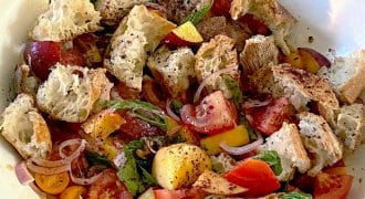 A bowl of panzanella salad with chunks of rustic bread, tomatoes, yellow cherry tomatoes, peach slices, red onion, fresh basil, and ground black pepper.