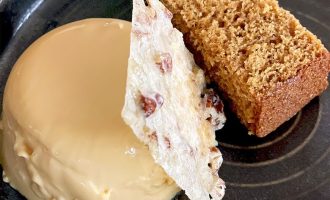 A round caramel-coloured dessert sits next to a slice of golden-brown cake, with a thin, textured tuile standing upright between them, all served on a dark ceramic plate.