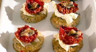 A white rectangular platter holds nine savoury appetisers made with a round biscuit base, topped with creamy cheese, roasted tomato halves, herbs, and small mushroom slices.