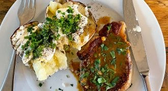A white plate with a baked potato topped with soured cream, chopped chives, and pepper, next to a pork chop with brown sauce and herbs. A fork and knife are placed on the plate. The table is wooden.