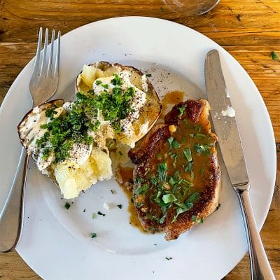 A white plate with a baked potato topped with soured cream, chopped chives, and pepper, next to a pork chop with brown sauce and herbs. A fork and knife are placed on the plate. The table is wooden.